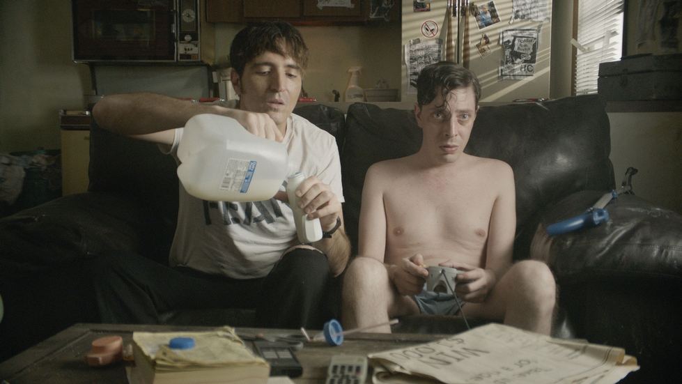 Two men in a living room sitting on a couch, one shirtless. The shirted man is pouring a glass of milk while the other is playing a video game